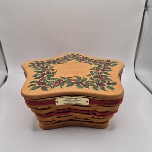 Puede incluir: Una cesta de madera en forma de estrella con acabado marrón claro y una banda decorativa roja trenzada. La tapa presenta un diseño pintado de una corona de acebo y bayas. Una placa de metal color latón indica Longaberger Christmas Collection 2001 Edition.