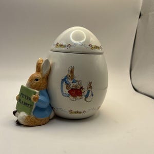 Può includere: Un barattolo di ceramica bianca a forma di uovo con coperchio, decorato con motivi floreali e illustrazioni di conigli. Una statuina di Peter Rabbit che tiene un libro è attaccata al lato. Il libro dice "Peter Rabbit".