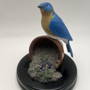 以下が含まれることがあります： 転倒した植木鉢にとまっている青い鳥のセラミックフィギュア。鳥は青い翼、茶色の胸、青い頭をしています。鉢は茶色で、土が紫の花の付いた台座にこぼれています。