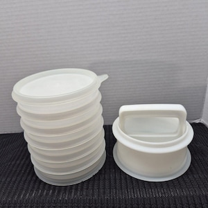 May include: A stack of eight translucent circular plastic lids with small pull tabs, positioned next to a white circular plastic press tool featuring a central handle. The items are constructed from smooth, semi-opaque material.