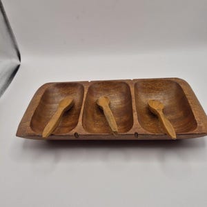 May include: A wooden serving tray with three compartments, each holding a small wooden spoon. The tray and spoons are a warm brown color, suggesting a natural wood finish. The rectangular tray has rounded edges.
