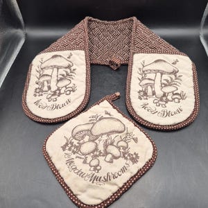 May include: A kitchen textile set with a mushroom design. The set includes a double oven glove and a square pot holder. The items are beige with a brown mushroom illustration and a brown polka dot trim. The text "Meadow Mushrooms" and "Wood Bloom" are visible.