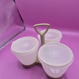 May include: A three-part serving dish with a handle. The dish has three white bowls with clear lids, set on a brown base. The handle is a light beige color. The background is a solid purple.