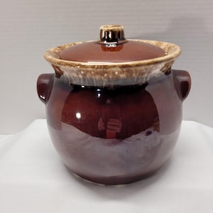 May include: A brown ceramic bean pot with a lid. The pot has two handles and a light brown rim. The lid has a small handle on top. The pot is a deep brown colour with a glossy finish.