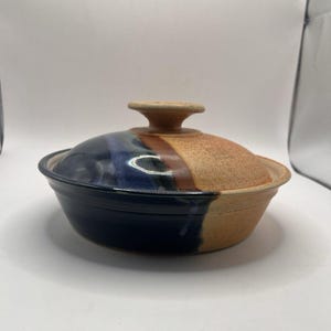 Könnte beinhalten: Eine runde Auflaufform aus Keramik mit Deckel und zweifarbiger Glasur. Eine Seite der Schüssel und des Deckels ist in tiefem Marineblau gehalten, während die andere Seite eine texturierte, erdige beigebraune Farbe aufweist.