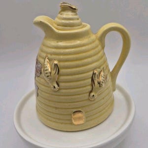 Puede incluir: Una cremera de cerámica amarilla con diseño de panal, detalles dorados y un asa pequeña. La cremera está sobre un platillo blanco. La cremera tiene un pico vertedor. La cremera mide aproximadamente 13 cm de alto.