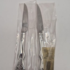 Può includere: Due coltelli color argento con lame seghettate, ciascuno in una custodia di plastica trasparente. I manici presentano un ornato disegno floreale. La confezione include il testo che recita "HERITAGE ANTI...Star Knives...MADE FAT."