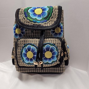 May include: Black backpack with embroidered blue and yellow floral design. The backpack features a flap closure, two front pockets, and gold-coloured hardware. The floral embroidery is detailed with green accents and the bag has a drawstring closure.