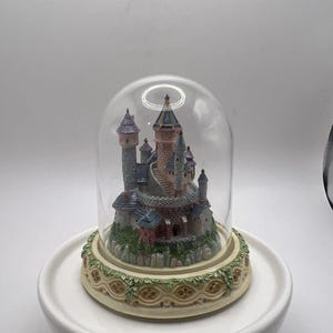 May include: A miniature castle figurine enclosed within a clear glass dome. The castle features multiple towers with blue and purple roofs, stone-textured walls, and a spiral staircase. It sits on a circular, cream-colored base with green vine detailing.
