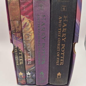 May include: A boxed set of four Harry Potter books. The spines are visible, showcasing titles like "Harry Potter and the Sorcerer's Stone" and "Harry Potter and the Goblet of Fire." The books are in a purple box.
