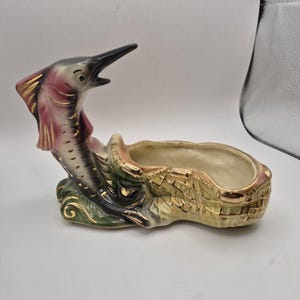 May include: A ceramic marlin figurine attached to a textured, basket-weave patterned bowl. The fish features a pink and grey body with gold-painted accents, while the bowl has a cream interior and a gold-trimmed rim, resting on a green base.
