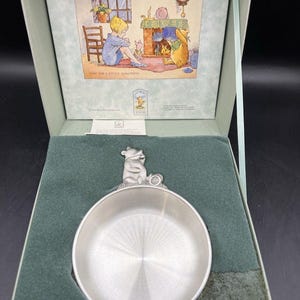 May include: A round pewter bowl featuring a sculpted Winnie the Pooh figure on the rim, presented in a light green box. The box lid displays the text Classic Winnie the Pooh Pewter Collection from Danforth Pewterers and an illustration.