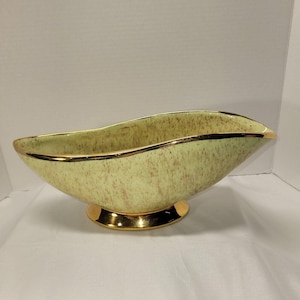 May include: A vintage, oblong-shaped ceramic bowl with a pale green speckled finish. The rim and base are trimmed with gold. The bowl is elevated on a gold-colored pedestal base. The interior of the bowl is a solid pale green color.