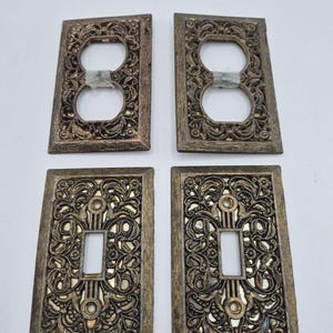 May include: A set of four rectangular wall plates with an ornate, raised floral and scrollwork pattern in a dark bronze finish. Two plates feature a double circular outlet opening, while the other two have a single rectangular switch opening.