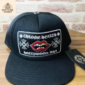 Könnte beinhalten: Schwarze Trucker-Cap mit Mesh-Rückseite und flachem Schirm. Auf der Vorderseite befindet sich ein rechteckiger Aufnäher mit dem Text "CHROME HEARTS HOLLYWOOD, USA" und einer Grafik roter Lippen mit einem Goldzahn.