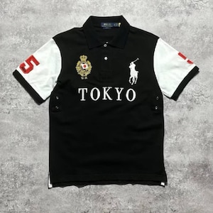May include: A black polo shirt with white sleeves, featuring the word TOKYO in white letters across the chest. It includes a white embroidered polo player logo, a gold crest with a Japanese flag, and a red number 5 on the right sleeve.