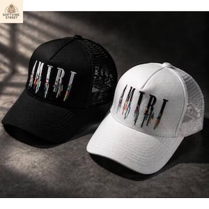 May include: A black trucker hat and a white trucker hat, both featuring a mesh back and the word AMIRI embroidered on the front in a stylized font with colorful, dripping paint-like details extending from the bottom of each letter.