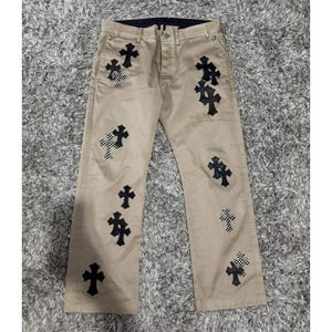 May include: Khaki-coloured trousers featuring numerous black cross appliqués. Some crosses have a chequered pattern. The trousers have a button and zip closure and belt loops. The trousers are laid flat on a grey carpet.
