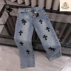 Puede incluir: Vaqueros de mezclilla azul claro con múltiples aplicaciones de cruces negras. Los vaqueros tienen un diseño clásico de cinco bolsillos y un corte de pierna recta. Una etiqueta y un llavero cuelgan del trabilla. Los vaqueros se exhiben sobre una superficie reflectante.