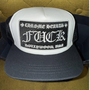 May include: A black and white trucker hat with a rectangular patch featuring the text "CHROME HEARTS FUCK HOLLYWOOD, USA" in a gothic font. The hat has a black brim and mesh back.