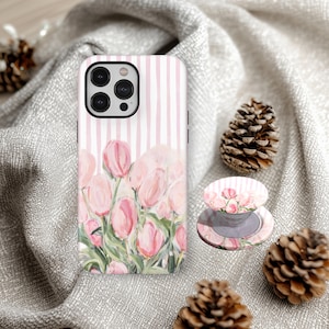 May include: A phone case and phone grip with a pink tulip floral design. The phone case has a vertical striped background with pink tulips. The phone grip has a matching design. The items are displayed on a grey fabric.