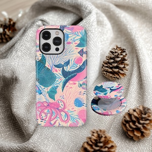 May include: A phone case and matching phone grip with a vibrant ocean design, including whales, fish, and an octopus. The case is predominantly pink, blue, and teal. The round phone grip mirrors the design. The items are displayed on a grey surface with pine cones.