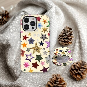 May include: A phone case and phone grip with a cream background and a pattern of colorful stars. The phone case is on a gray fabric surface with pine cones.