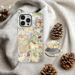 Könnte beinhalten: Eine Handyhülle und ein Handyhalter mit einem skurrilen Waldmotiv mit Hasen, Enten, Pilzen und Blumen. Die Hülle ist hellgrün mit einem sich wiederholenden Muster. Der Handyhalter ist rund und hat ein passendes Design. Kiefernzapfen sind verstreut.