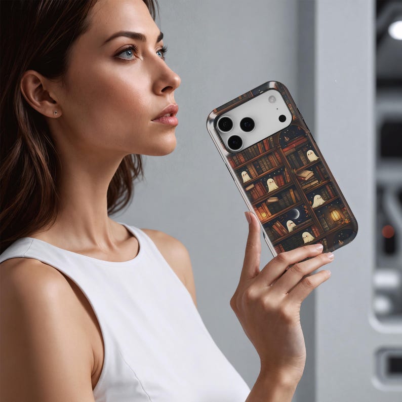 Puede incluir: Funda de tel&eacute;fono inteligente con un dise&ntilde;o de estanter&iacute;a llena de libros, fantasmas y una luna creciente. La funda tiene un fondo oscuro con iluminaci&oacute;n c&aacute;lida, creando una est&eacute;tica acogedora y espeluznante. La funda la sostiene una persona.