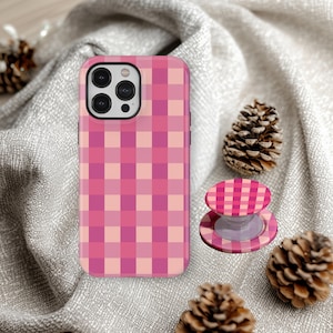 May include: A rectangular smartphone case and a matching circular collapsible phone grip, both featuring a repeating pink, light pink, and magenta checkered pattern. The phone case has a cutout for a triple-lens camera system on the back.