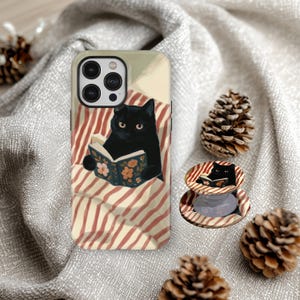 Puede incluir: Una funda de teléfono y un agarre para teléfono con un gato negro leyendo un libro con ilustraciones florales. La funda tiene un fondo beige con una ilustración de un gato negro en una cama con rayas rojas y blancas. El agarre tiene un diseño a juego.