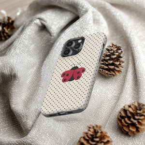 May include: A phone case with a cream-colored background and a pattern of small black dots. A red and black ladybug design is centered on the case. The phone case is surrounded by pine cones and a gray fabric.