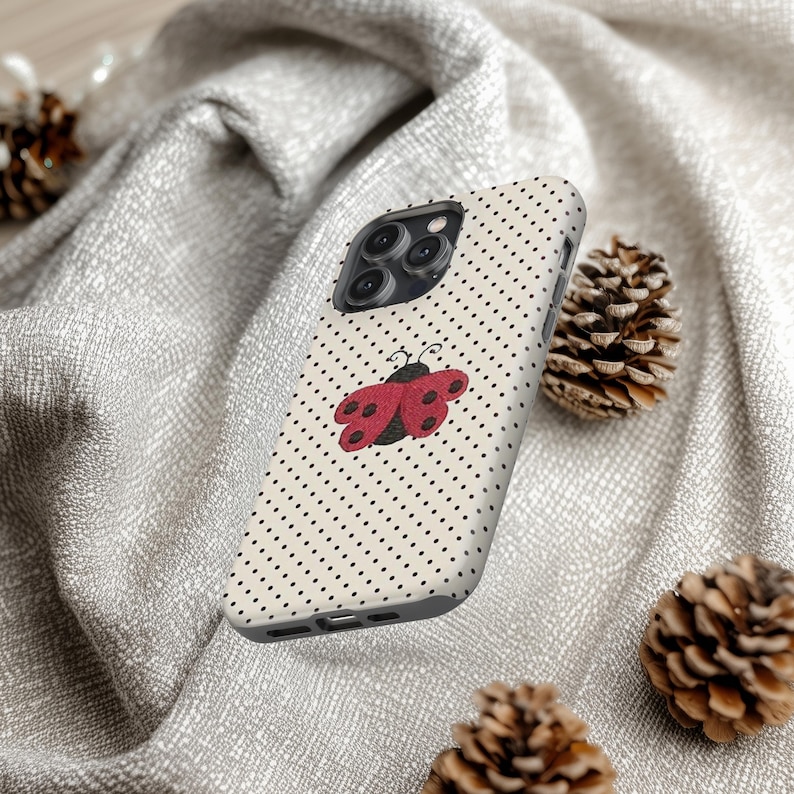 May include: A phone case with a cream-colored background and a pattern of small black dots. A red and black ladybug design is centered on the case. The phone case is surrounded by pine cones and a gray fabric.
