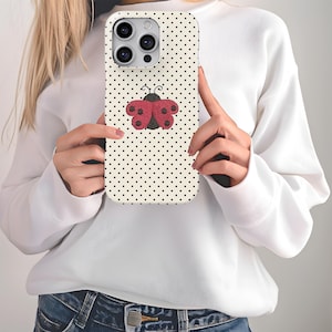 May include: A phone case with a cream background covered in small black polka dots. A red and black ladybug design is centered on the case. The phone case is held by a person wearing a white sweater and blue jeans.