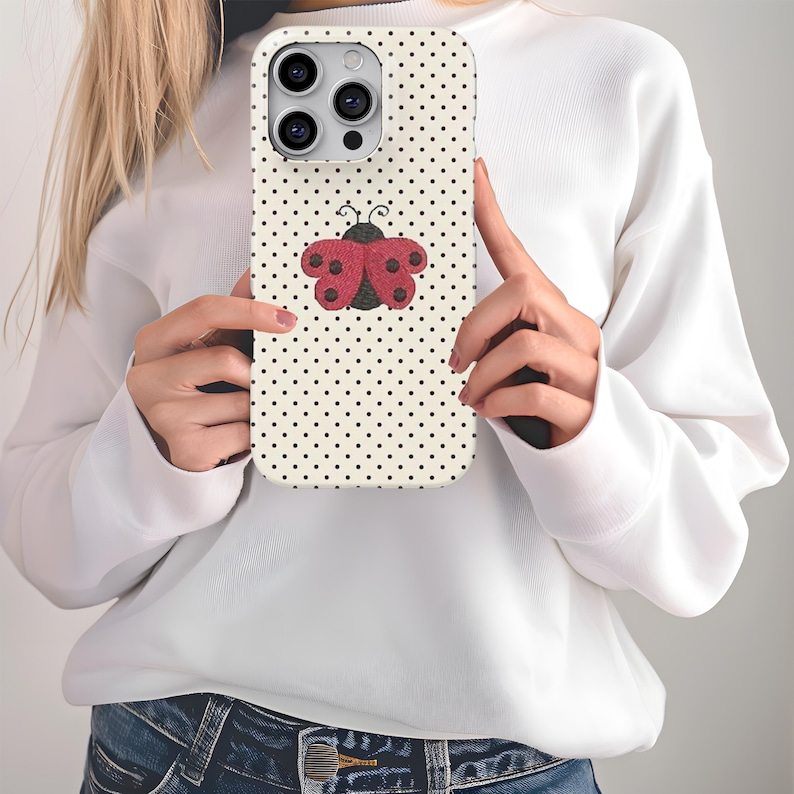 May include: A phone case with a cream background covered in small black polka dots. A red and black ladybug design is centered on the case. The phone case is held by a person wearing a white sweater and blue jeans.
