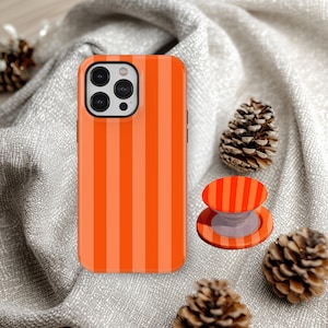 May include: Orange and light orange striped phone case and matching pop socket. The phone case is on a gray surface with pinecones.