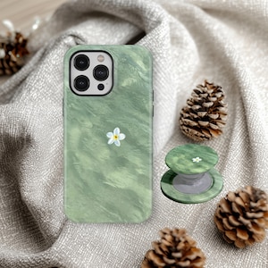 May include: Green phone case and matching PopSocket with a white flower design. The case has a marbled texture. The PopSocket is round and has a white flower in the center. The background includes pine cones and a gray fabric.