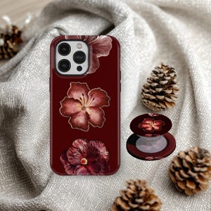Puede incluir: Una funda de teléfono burdeos y un soporte emergente a juego con un diseño floral. La funda tiene tres ilustraciones de flores de hibisco. El soporte emergente es redondo con un diseño floral similar. Hay piñas en el fondo.