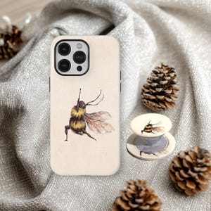 Könnte beinhalten: Beige Handyhülle und passender Handyhalter mit einer Aquarellillustration einer Hummel. Die Biene wird im Flug mit ausgebreiteten Flügeln dargestellt. Das Set befindet sich auf einem grauen Stoffhintergrund mit Tannenzapfen.