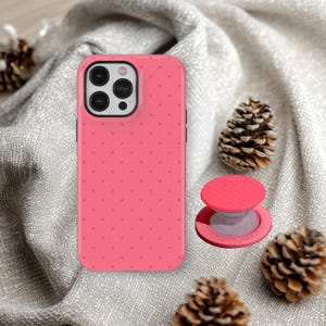 Può includere: Una cover per smartphone rosa brillante con un motivo ripetuto di piccoli punti rosa scuro, abbinata a un supporto circolare pieghevole per telefono con parte superiore rosa e base grigio chiaro, adatta a un dispositivo con tripla fotocamera.