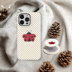 May include: A cream-colored phone case with black polka dots and a red ladybug design. A matching phone grip is also visible. The case and grip are on a gray fabric surface, with pinecones in the background.