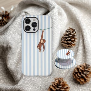 May include: A phone case and phone grip featuring a cartoon monkey hanging from a green line. The case has a blue and white striped pattern. The phone grip has the same design. The background includes pinecones.