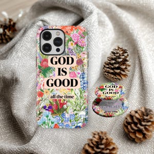May include: A phone case and pop socket with a floral design and the text "GOD IS GOOD all the time". The case and pop socket have a colourful flower pattern with pink, red, blue, and green shades. Two pine cones are also in the image.