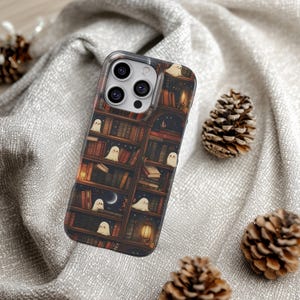 Puede incluir: Una funda transparente para tel&eacute;fono con un dise&ntilde;o de estanter&iacute;a y peque&ntilde;as ilustraciones de fantasmas blancos. La estanter&iacute;a tiene una paleta de colores c&aacute;lidos con libros, velas y una escena de cielo nocturno. La funda est&aacute; rodeada de pi&ntilde;as sobre una superficie gris texturizada.
