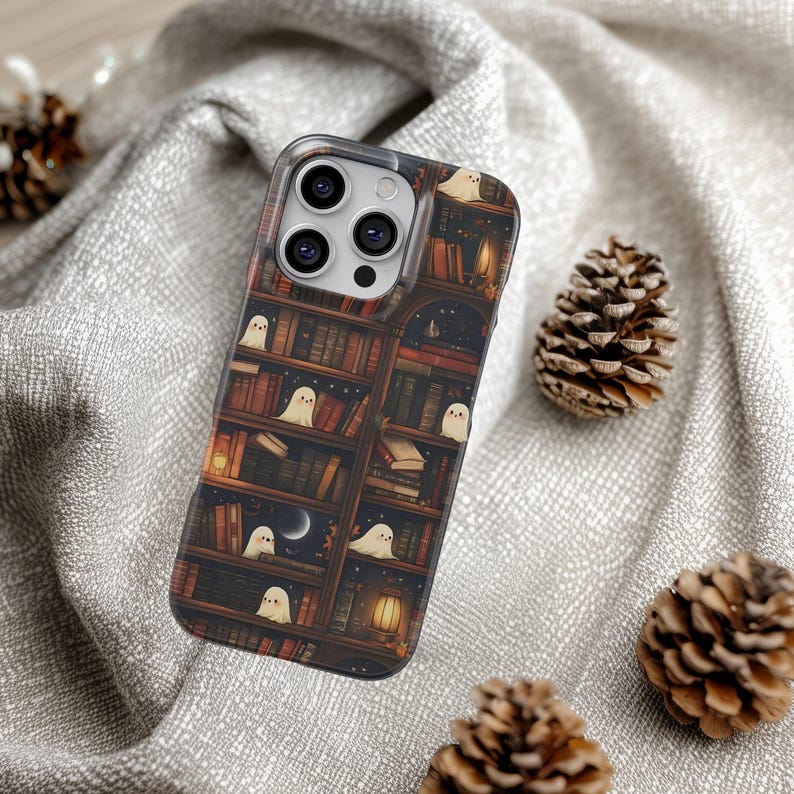 Puede incluir: Una funda transparente para tel&eacute;fono con un dise&ntilde;o de estanter&iacute;a y peque&ntilde;as ilustraciones de fantasmas blancos. La estanter&iacute;a tiene una paleta de colores c&aacute;lidos con libros, velas y una escena de cielo nocturno. La funda est&aacute; rodeada de pi&ntilde;as sobre una superficie gris texturizada.
