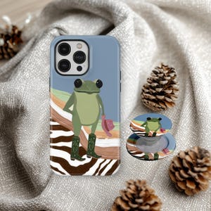 May include: A light blue phone case and matching pop socket featuring a cartoon frog wearing cowboy boots and a pink hat. The case has a colorful landscape design with a zebra print base.