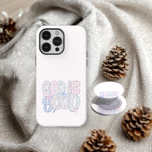 May include: White phone case with a polka dot pattern and the words "GOD IS GOOD" in pastel colors. A matching phone grip is also visible. The phone case and grip are on a gray fabric surface, with pine cones.