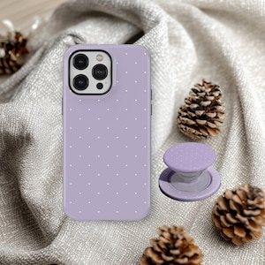 May include: A lavender phone case and matching pop socket with a white polka dot pattern. The phone case is shown with a black camera lens and a silver rim. The pop socket is next to the phone case. The background is a gray fabric.