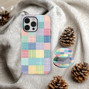 Puede incluir: Una funda de teléfono y un soporte a juego con un diseño de patchwork pastel. La funda y el soporte presentan una cuadrícula de cuadrados coloridos en tonos rosa, azul, verde, amarillo y morado. La funda del teléfono tiene un módulo de cámara negro.