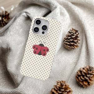 May include: A phone case with a cream-colored background and black polka dots. A red and black ladybug design is centered on the case. The case has a camera cutout and is surrounded by pine cones.